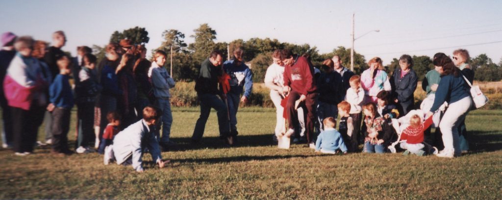Picture of four men holding two shovels about to dig into the ground. There is a crowd of people around watching.