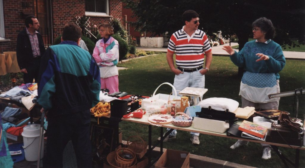 Picture of a man with a cane looking at items for sale at a yard sale table. In the back are the sellers standing and talking.